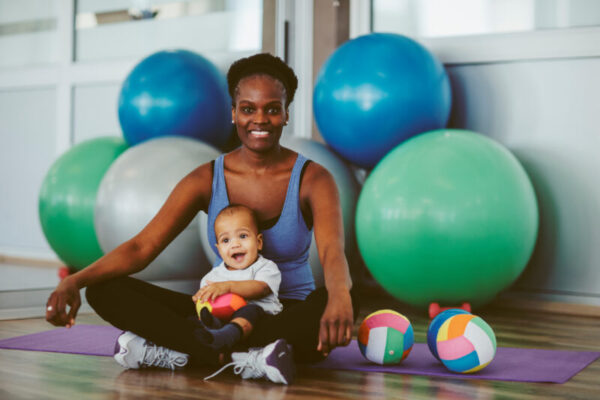 Mother with her baby boy in the gym sitting on the floor after exercising. They are looking at camera and smiling. Mother is from South African, living in Europe. Her cute son is mixed race.